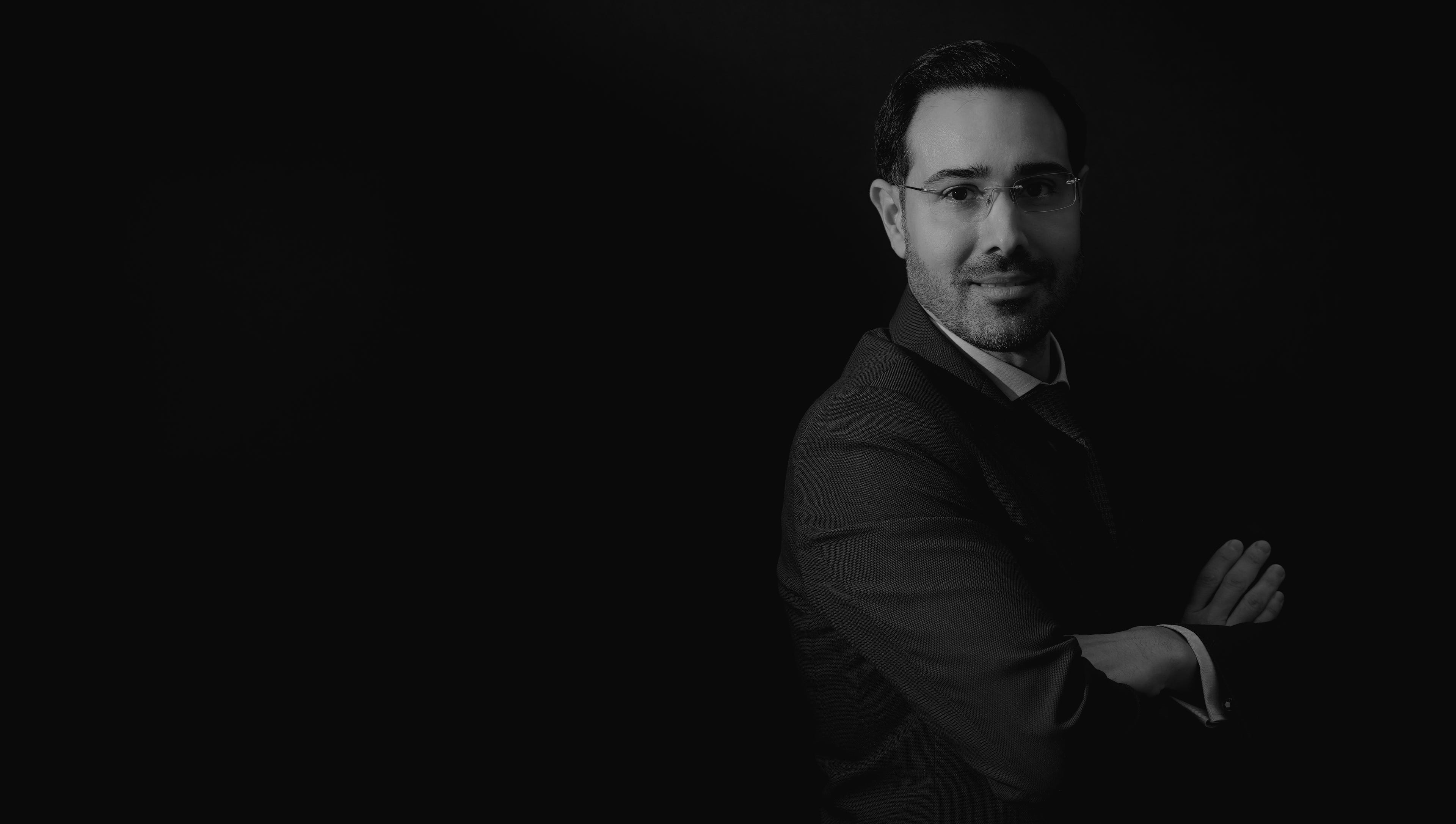 Dr. Nabil Fakih Photo in glasses and a suit stands with arms crossed against a dark background.